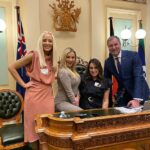 De izquierda a derecha: Tamika Smith, Brooke Henley-Smith y Lauren Duitsch posan con el parlamentario John-Paul Langbroek frente a la silla del presidente del Parlamento de Queensland.