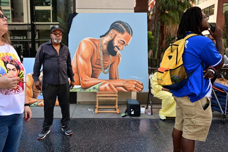 Artist Mohammed Mubarak stands beside his artwork of the late US rapper Nipsey Hussle as people gather for Hussle's posthumous Hollywood Walk of Fame Star ceremony in Hollywood, California, on August 15, 2022. - Slain rapper Nipsey Hussle, who was shot in Los Angeles three years ago, was honored with a star on Hollywood's Walk of Fame on what would have been his 37th birthday. (Photo by Frederic J. BROWN / AFP) (Photo by FREDERIC J. BROWN/AFP via Getty Images)