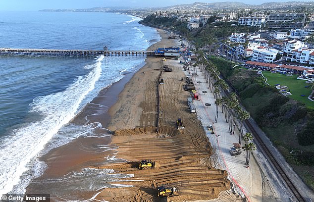 Los trabajadores mueven arena fresca entregada en barcaza a la playa pública principal durante un proyecto de reposición de arena a lo largo de la costa erosionada el 21 de mayo de 2024 en San Clemente, California.