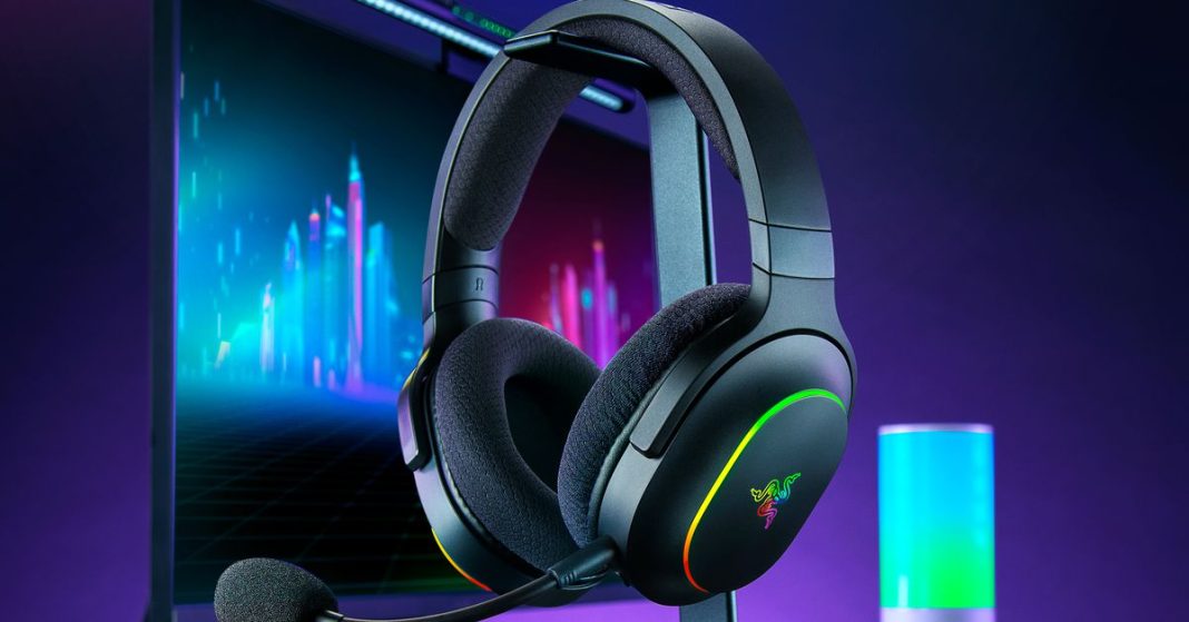 The Razer Barracuda X Chroma headphones hanging on a stand in front of a glowing gaming rig.