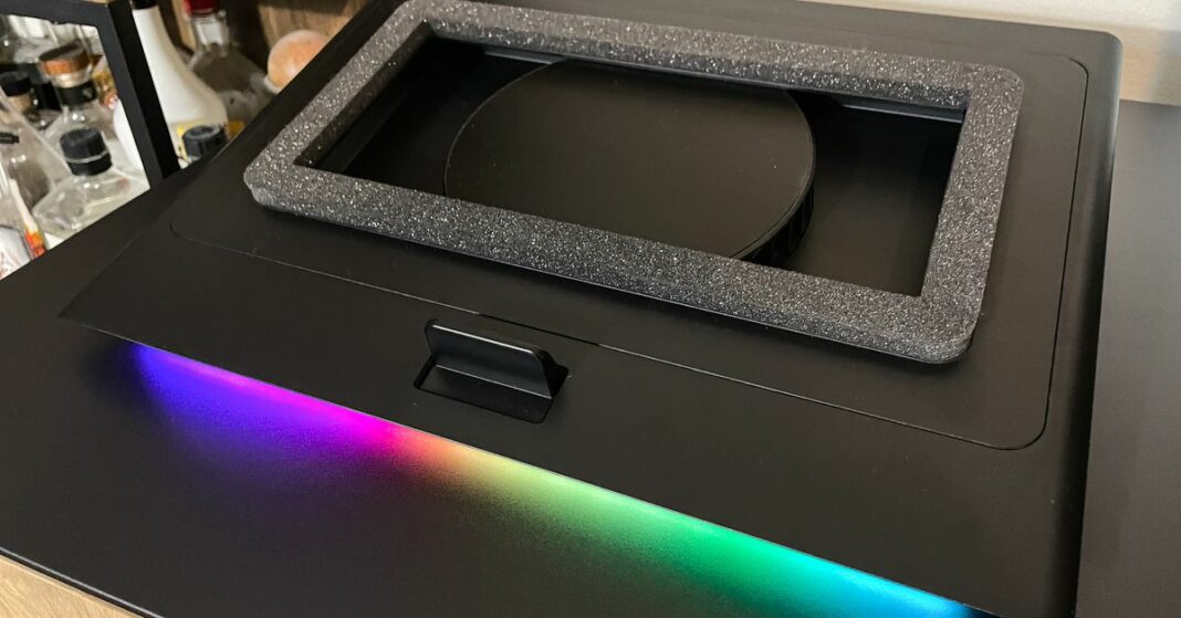 A laptop stand with a rectangular cutout. The cutout has a foam seal and a cooling fan inside, and the stand casts an RGB glow onto the surface below.