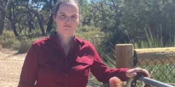 Mujer con camisa roja de pie junto a una puerta en un entorno natural.