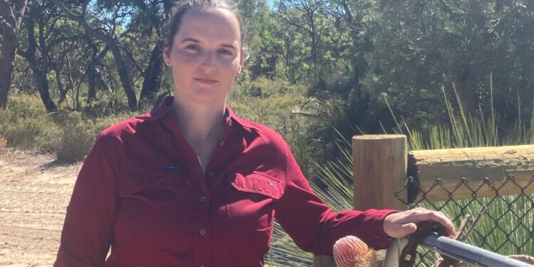 Mujer con camisa roja de pie junto a una puerta en un entorno natural.