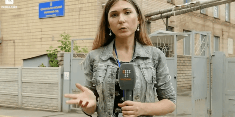 Reportero sosteniendo un micrófono frente a un edificio.