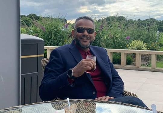 Hombre sentado al aire libre en una mesa, disfrutando de una bebida.