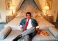 Bob Guccione sentado en una cama con una chaqueta de mezclilla y corbata roja, sosteniendo revistas.