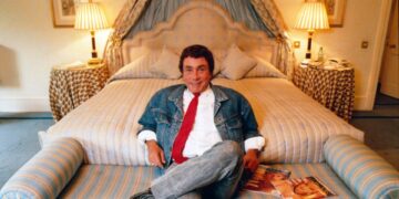 Bob Guccione sentado en una cama con una chaqueta de mezclilla y corbata roja, sosteniendo revistas.