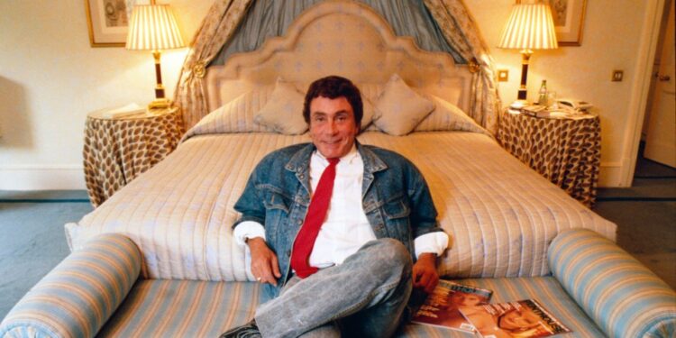 Bob Guccione sentado en una cama con una chaqueta de mezclilla y corbata roja, sosteniendo revistas.