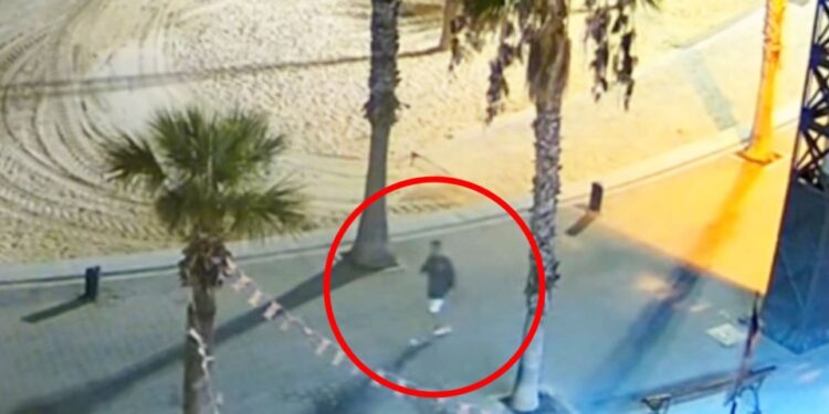 Imagen de CCTV de una persona que camina en un área pavimentada cerca de una playa por la noche.