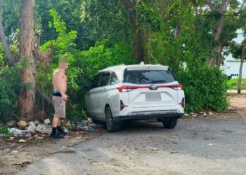 Un hombre sin camisa se encuentra cerca de un Toyota Veloz blanco en un estacionamiento.
