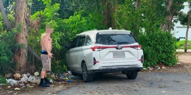Un hombre sin camisa se encuentra cerca de un Toyota Veloz blanco en un estacionamiento.