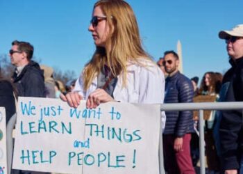 Un científico en su bata de laboratorio tiene un letrero que lee "¡Solo queremos aprender cosas y ayudar a las personas!" Durante una demostración al aire libre