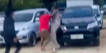 Un hombre con una camisa roja que pelea con otro hombre cerca de autos estacionados.