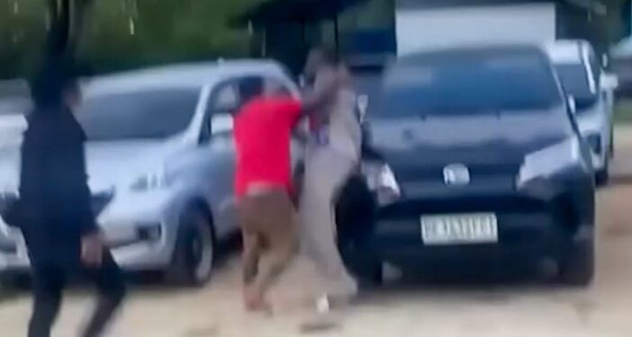 Un hombre con una camisa roja que pelea con otro hombre cerca de autos estacionados.