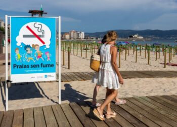 Señal de playa que prohíbe fumar, con gente caminando en un paseo marítimo.