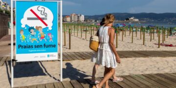 Señal de playa que prohíbe fumar, con gente caminando en un paseo marítimo.
