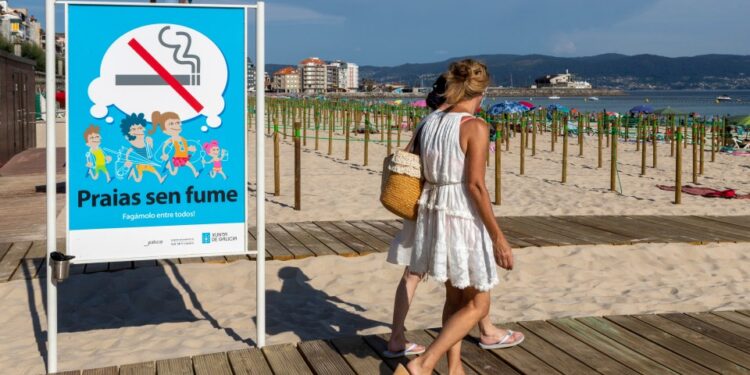 Señal de playa que prohíbe fumar, con gente caminando en un paseo marítimo.