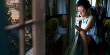 Un niño pequeño con discapacidades del Agente Orange mira afuera mientras se sienta en un alféizar