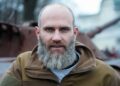 Retrato de un hombre con una larga barba gris, con una chaqueta bronceada, cerca de vehículos militares destruidos.