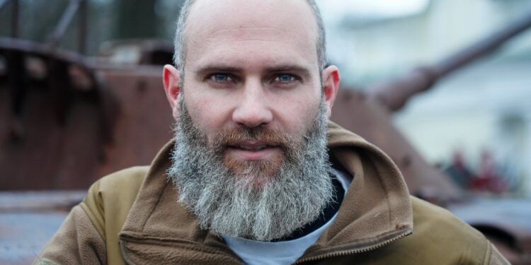 Retrato de un hombre con una larga barba gris, con una chaqueta bronceada, cerca de vehículos militares destruidos.