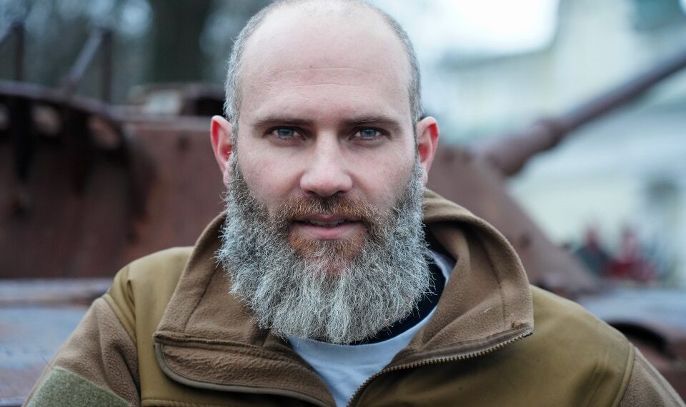 Retrato de un hombre con una larga barba gris, con una chaqueta bronceada, cerca de vehículos militares destruidos.