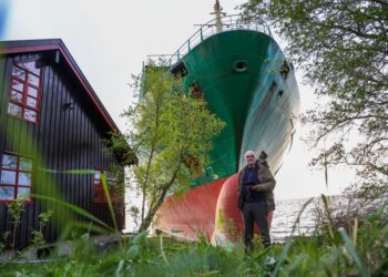 Hombre parado frente a la casa con un barco a tierra en el fondo.