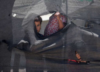 Tres bebés palestinos miran a través de agujeros desde detrás de una lona desgarrada