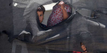 Tres bebés palestinos miran a través de agujeros desde detrás de una lona desgarrada