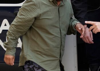 Un hombre esposado con una camisa verde y pantalones cortos estampados es escoltado en un vehículo policial.