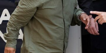 Un hombre esposado con una camisa verde y pantalones cortos estampados es escoltado en un vehículo policial.