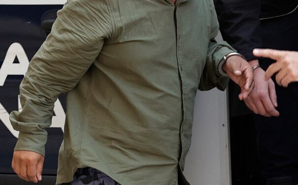 Un hombre esposado con una camisa verde y pantalones cortos estampados es escoltado en un vehículo policial.