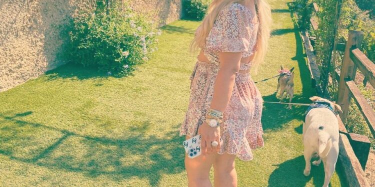 Mujer con un vestido rosa y sombrero caminando dos perros en un camino cubierto de hierba.