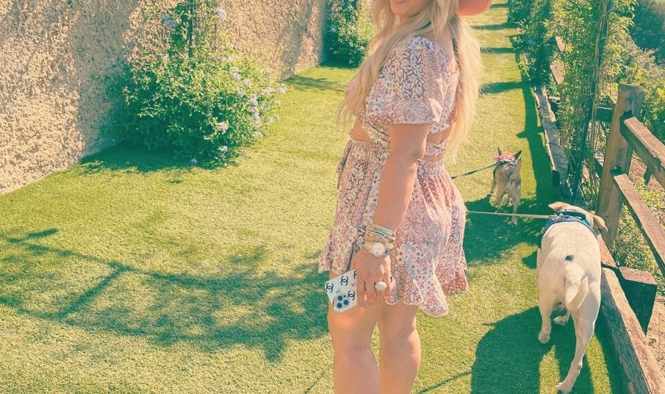 Mujer con un vestido rosa y sombrero caminando dos perros en un camino cubierto de hierba.