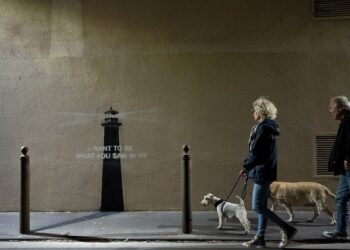 Dos personas pasan a sus perros  allá de una pared con una plantilla de faro y las palabras "Quiero ser lo que viste en mí."