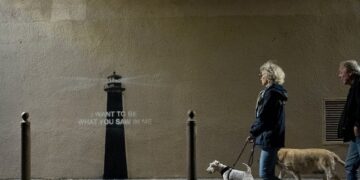 Dos personas pasan a sus perros  allá de una pared con una plantilla de faro y las palabras "Quiero ser lo que viste en mí."