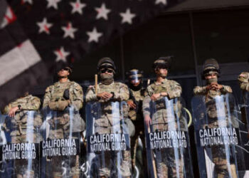 Las tropas de la Guardia Nacional de California se forman en formación mientras un manifestante agita una interpretación en blanco y negro manchado de sangre de la bandera estadounidense en primer plano