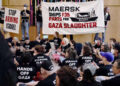 Personas sentadas en el suelo con camisas negras con texto blanco que lee "Manos fuera de Gaza", "Deja de hambre de Gaza" y "Terminar con el asedio" aplaudir y animar mientras otros sostienen las pancartas leyendo "Maersk envía piezas F35 para la matanza de Gaza" y "Deja de armar a Israel" Durante un rally interior