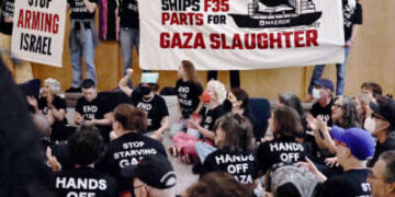 Personas sentadas en el suelo con camisas negras con texto blanco que lee "Manos fuera de Gaza", "Deja de hambre de Gaza" y "Terminar con el asedio" aplaudir y animar mientras otros sostienen las pancartas leyendo "Maersk envía piezas F35 para la matanza de Gaza" y "Deja de armar a Israel" Durante un rally interior