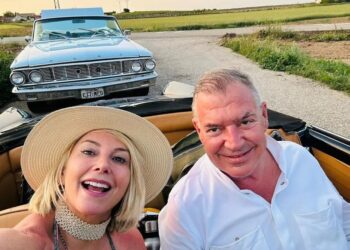 Un hombre y una mujer en un automóvil convertible, con un auto clásico detrás de ellos.