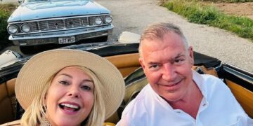 Un hombre y una mujer en un automóvil convertible, con un auto clásico detrás de ellos.