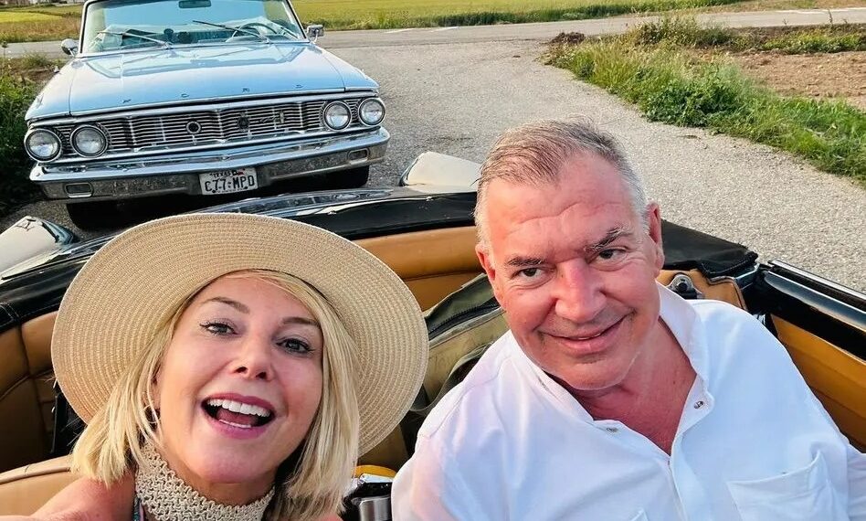 Un hombre y una mujer en un automóvil convertible, con un auto clásico detrás de ellos.