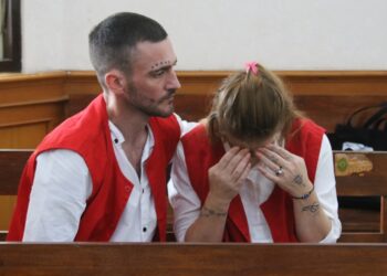 Un hombre y una mujer en chalecos rojos se sientan en la corte; La mujer está llorando.