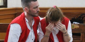 Un hombre y una mujer en chalecos rojos se sientan en la corte; La mujer está llorando.