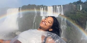 Mujer en un poncho de plástico sonriendo frente a una cascada con un arco iris.