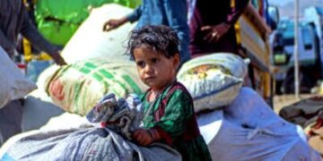 Un niño pequeño se agarra a una bolsa más grande que ella como hombres detrás de su carga un camión