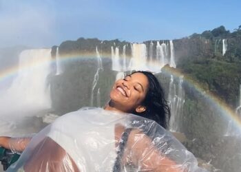 Mujer en un poncho de plástico sonriendo frente a una cascada con un arco iris.