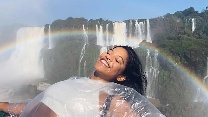 Mujer en un poncho de plástico sonriendo frente a una cascada con un arco iris.