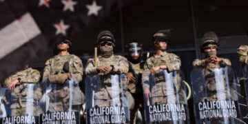 Las tropas de la Guardia Nacional de California se forman en formación mientras un manifestante agita una interpretación en blanco y negro manchado de sangre de la bandera estadounidense en primer plano
