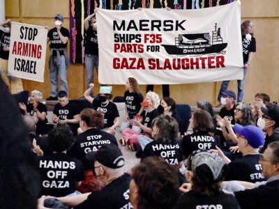 Personas sentadas en el suelo con camisas negras con texto blanco que lee "Manos fuera de Gaza", "Deja de hambre de Gaza" y "Terminar con el asedio" aplaudir y animar mientras otros sostienen las pancartas leyendo "Maersk envía piezas F35 para la matanza de Gaza" y "Deja de armar a Israel" Durante un rally interior