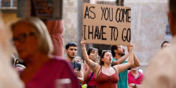 Manifestante sosteniendo un letrero que dice "Como vengo, tengo que irme" Frente a los turistas.